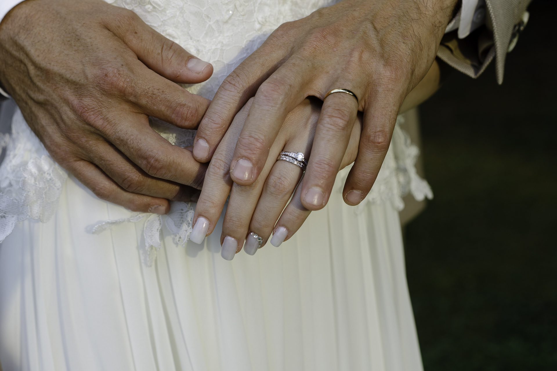 Deux mains se touchent, montrant leurs alliances fraîchement installées à leur doigts. En fond, la robe de la mariée indique que cette photo a été prise lors du mariage.