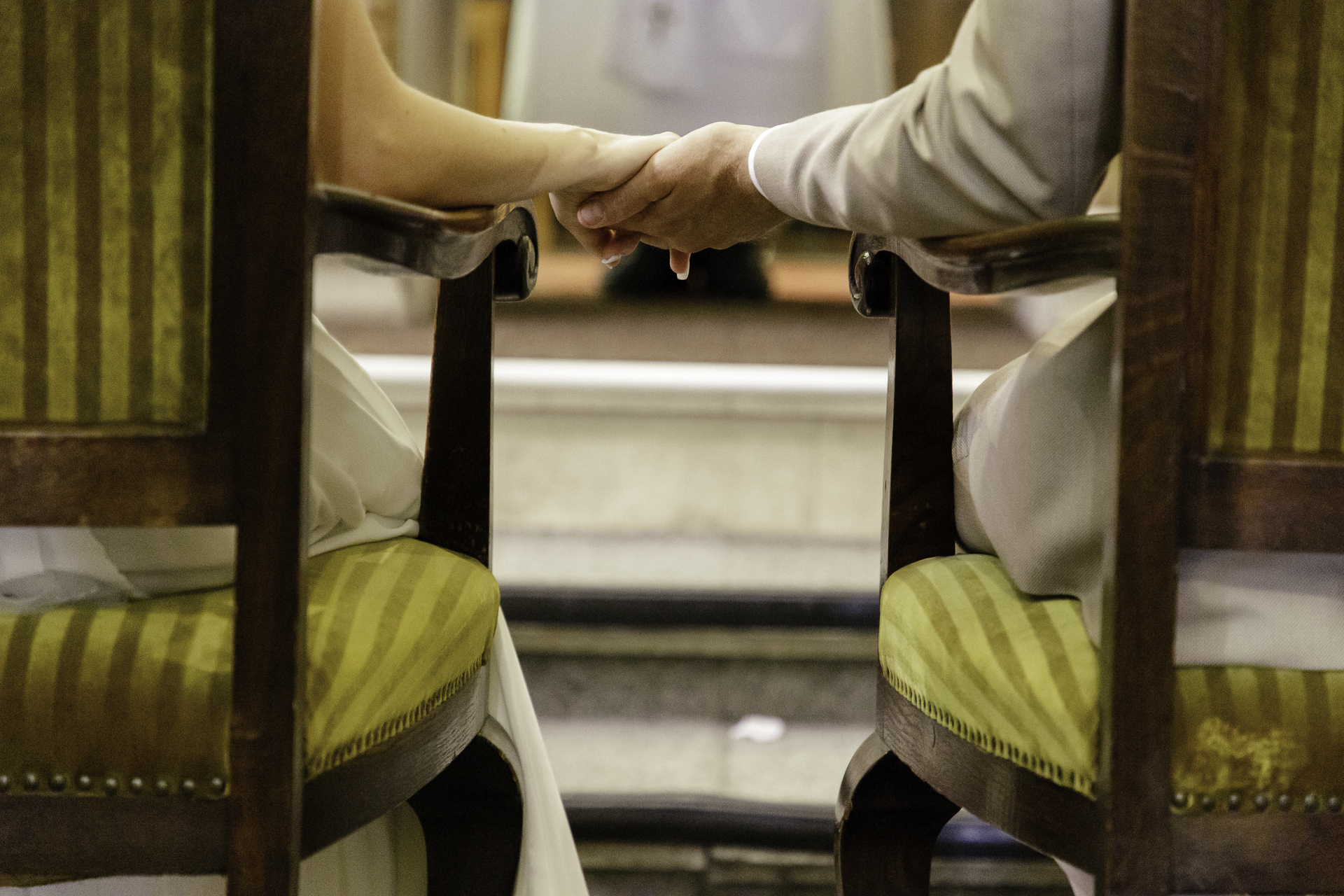 Photo de détail lors de la cérémonie religieuse. Les mariés, de dos, sont installés sur des fauteuils, qui se trouvent de chaque coté de la photographie. En son centre, leur main se rencontre pour symboliser leur envie de partager ce moment ensemble.